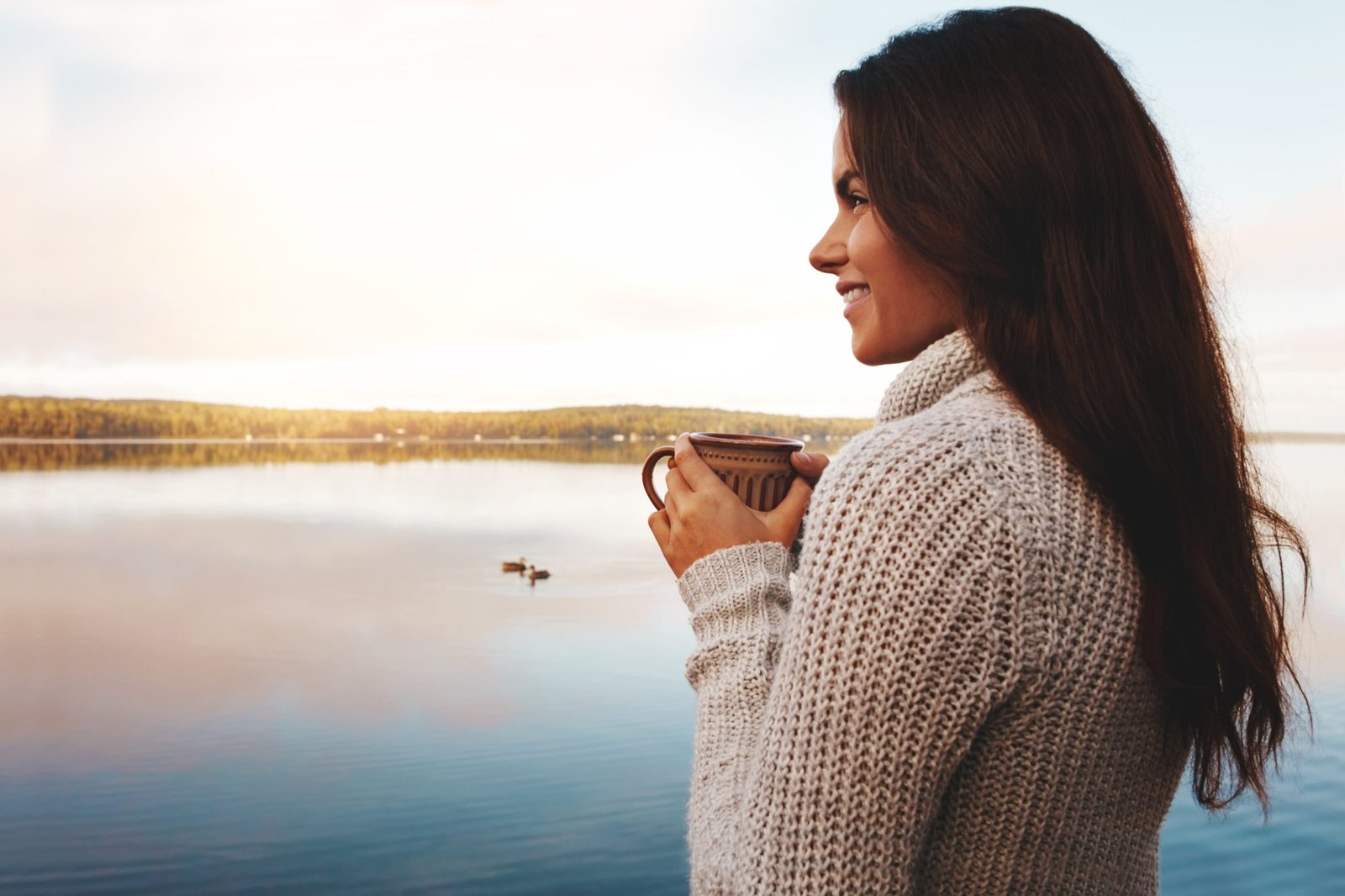 smile Outdoor, smile and woman with coffee in river, sunrise and happy in nature for vacation and morning. Break, writer and girl thinking of inspiration in countryside of Finland with peace and lake.