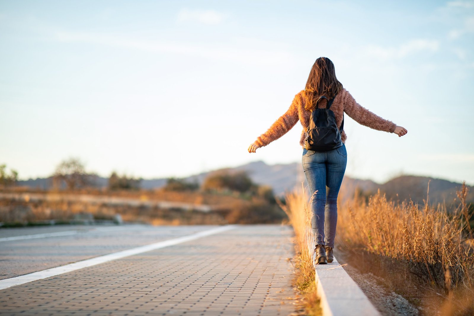 Beautiful woman walking and balancing on street curb or curbstone during sunset with copy space